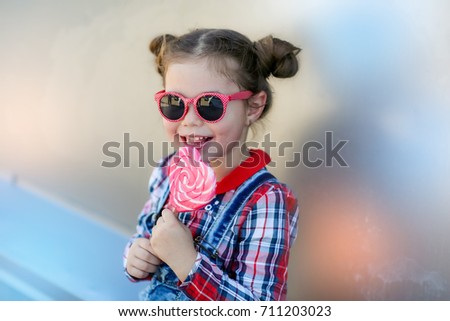 Stylish baby girl in sunglasses, with a haircut in a jeans dress with candy in the shape of a heart