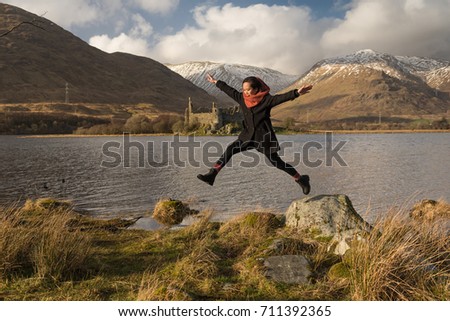 A girl wearing black clothes and a scarf from a rock on a lakeside with the Kilchurn Castle across the Loch Awe in the distance in the Scottish Highlands, UK, with snowy mountains on the background.