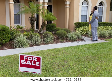 A happy African American man and woman couple house hunting outside a large house with a For Sale sign