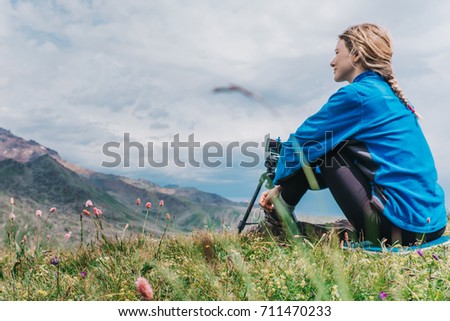 Girl sits on a tourist seat and looking on a tall mountain with mobile phone on tripod