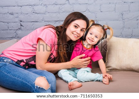 mother and daughter of two years listening to music with headphones on their head. Sit on the couch in the room against a blue brick wall. Embrace and rejoice and smile
