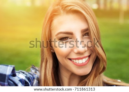 Selfie of gorgeous young woman in park. Closeup self portrait of beautiful blonde girl with big toothy smile. Retouched, vibrant colors.