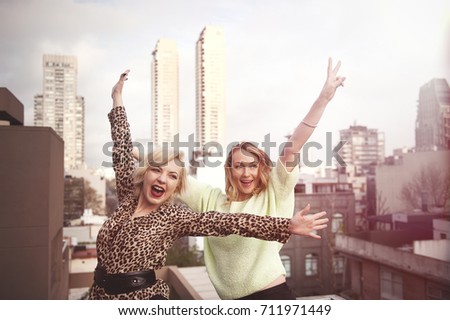two blond women friends standing on the balcony with hands up and laughing