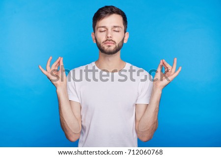 Attractive European hipster with peaceful expression meditating, holding his hands in mudra gesture, keeping his eyes closed while practicing yoga in office, trying to calm down and relax on break. 