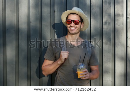 Horizontal image of handsome Caucasian guy pictured on grey wooden background in rural environment wearing casual clothes and hat, looking rightwards through sunglasses, discovering interesting places