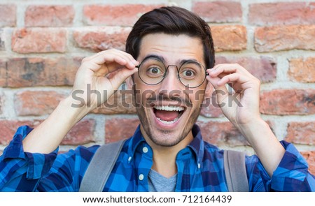 Horizontal portrait of young handsome guy pictured isolated on red brick wall holding round spectacles of bridge of nose in strong amazement as if having seen unexpectedly beneficial offer or chance