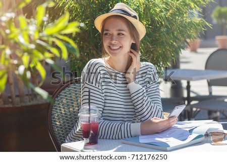 Horizontal picture of young beautiful European woman in beige hat with ribbon looking aside with curiosity to street while using cellphone for communication and spending afternoon in outdoor cafe