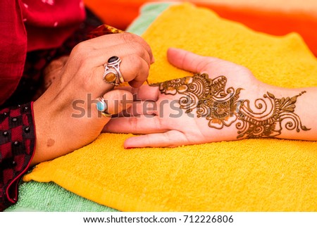 Mehndi artist carefully painting an intricate design using henna, on the hand and forearm of young Indian woman before a Vivaah-Indian Wedding. Arm resting on bright towels.