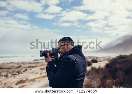 Side view shot of young photographer photographing outdoors. Man taking a photos with dslr camera outdoors. Photographer taking travel nature photography.