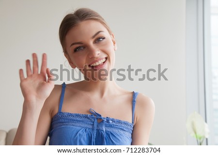 Smiling teen girl waving hand looking at web camera, happy young lady making video call at home, greeting online by webcam or recording videoblog, saying hello starting broadcast, head shot portrait