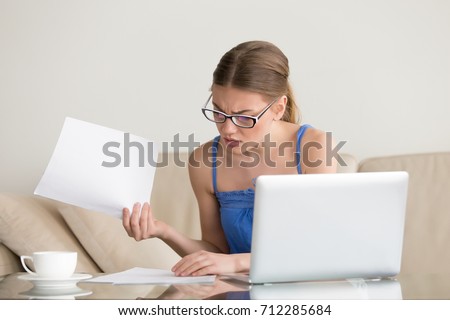 Confused worried woman looks through papers sitting on sofa at home, having problem with documents, found mistake in account statements, frustrated by huge domestic bills, reads letter with bad news Royalty-Free Stock Photo #712285684