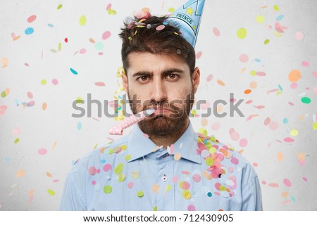 Unhappy birthday guy with stubble feeling sad and disappointed because nobody came to celebrate his anniversary, blowing party horn all alone, confetti flying around him. People and celebration Royalty-Free Stock Photo #712430905