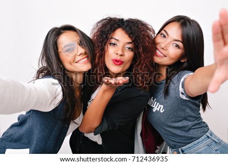 Inspired latin girl in glasses taking photo of herself while fooling around with university friends. Romantic curly black woman sending air kiss for selfie with charming ladies.