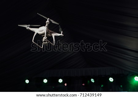 White quadrocopter fly inside the party-tent . Dark background, club light and soffits. Take photos and record footage.