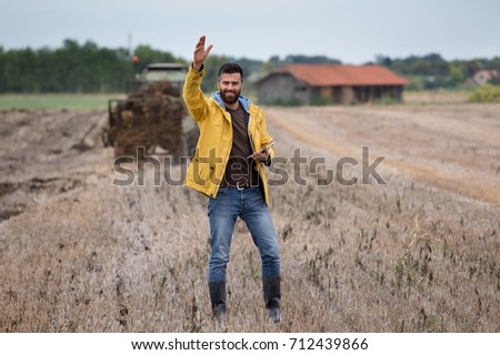 Young attractive man with beard holding tablet in field with tractor working in background