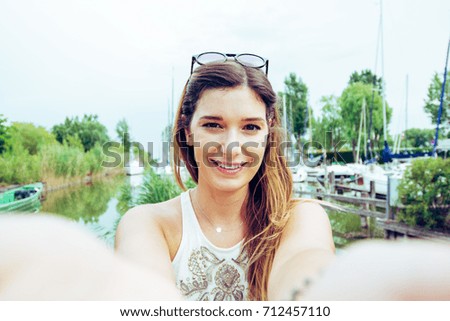 Beautiful young woman making a selfie near port and smiling in camera. Brunette girl spending good time and sharing on social network her self photo with smartphone. Concept of social addicted people.