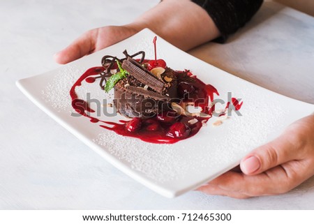 Customer holds chocolate cake with cherry jam, decorated by sweet red berry, almond and mint. Delicious dessert serving in restaurant