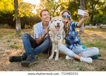 young beautiful couple sitting in park, smiling, happy friends together, casual denim outfit, summer style, making selfie photo with a husky dog, having good time