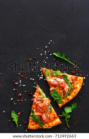 Slices of pizza with prosciutto and rocket salad with spices on black background, copy space, top view