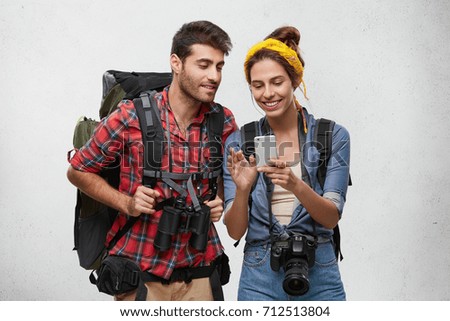 Two best friends preparing for trip in mountains, taking camera and binoculars with them, looking in smart phone, watching photos which they made during their summer vacations having happy look