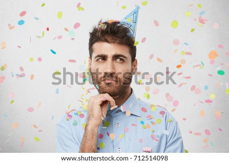 Studio shot of thoughtful pensive young Caucasian man wearing holiday cap and formal shirt looking sideways, having mysterious expression, holding hand on chin. Party, fun, celebration and birthday