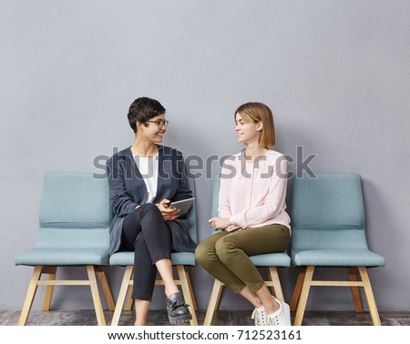 Lively conversation in queue. Two unemployed attractive young women in strict clothing talking to each other at HR department and discussing their resumes while waiting their turn for job interview Royalty-Free Stock Photo #712523161