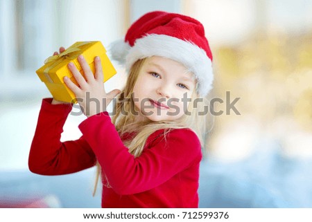 Adorable little girl wearing Santa hat opening a giftbox on Christmas morning. Celebrating Xmas at home.