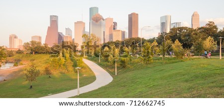 Panorama of downtown Houston, Texas, USA at sunset from Eleanor Tinsley Park. Grassy green park lawn, Buffalo Bayou river, curved pathway with people walking, biking, exercising and modern skylines.