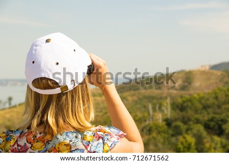 A girl in a blouse with flowers and a white cap looks through binoculars against a clear blue sky and green mountains on a sunny summer day.