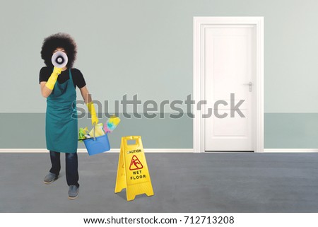 Picture of Afro maid shouting with a megaphone while holding a bucket and standing near a board of wet floor sign