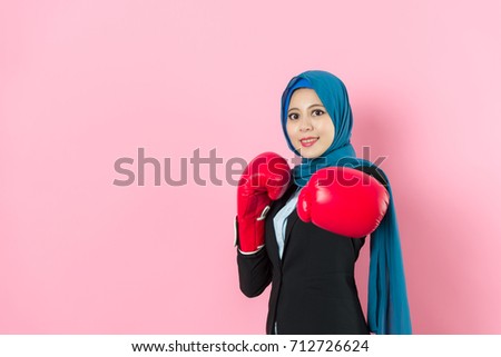 confident pretty muslim business woman face to camera smiling making hitting posture when she wearing boxing gloves standing in pink background. Royalty-Free Stock Photo #712726624