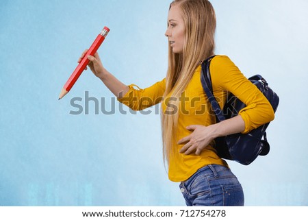 Happy young blonde teenage girl going to school or college wearing backpack and holding big pencil.