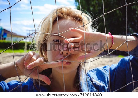 young woman in jeans clothes outdoors. portrait of a girl with freckles on her face, against the background of a football gate grid, on a sunny summer day. hands and fingers