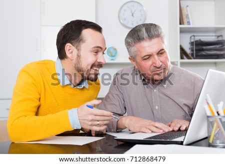 Smiling adult and young men look interesting on Internet on laptop at desk at ofice