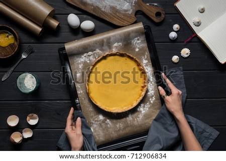 Top view shot of female hands holding a baking tray with freshly baked pumpkin pie over a wooden table covered with cooking ingridients