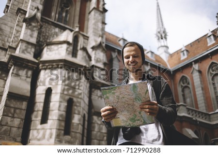 The picture of a traveller that is standing near the church. He is holding the map in his hands. He has found the place where he wants to go. This guy looks happy. Cut view