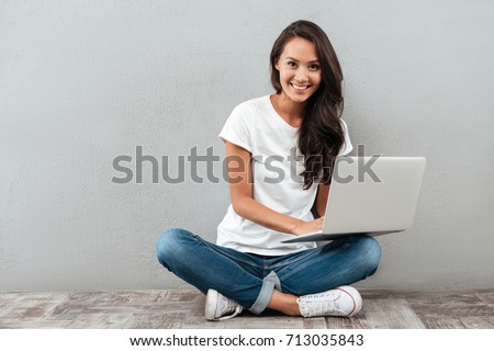 Smiling asian woman working on laptop computer while sitting on the floor with legs crossed isolated over gray background