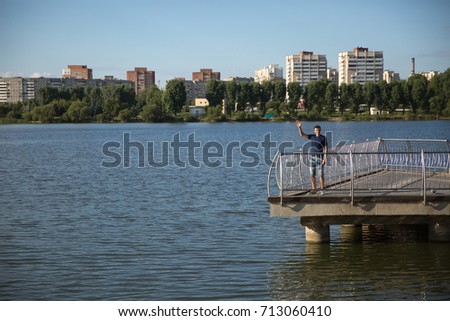 The man on the pier makes a welcome gesture with his hand . In a blue tank top against the backdrop of the city . On the river bank