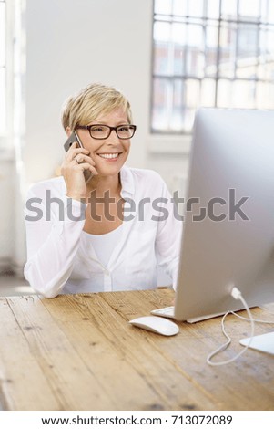 Businesswoman talking on a mobile in the office as she sits at her computer with a pleased happy smile