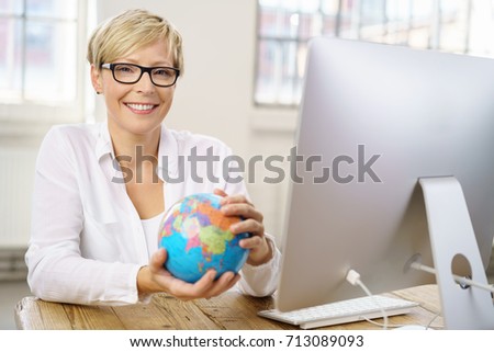 Smiling attractive young businesswoman sitting holding a globe at her desk in the office in a conceptual image