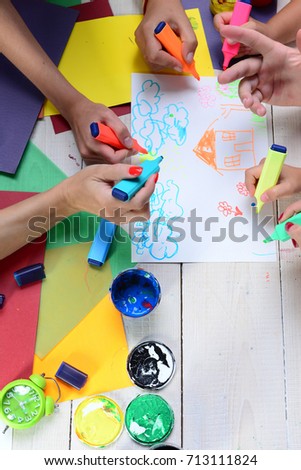 Hands hold colorful markers and draw. Artists wooden table with paints and colored paper. Art and idea concept. Markers in male and female hands draw kids illustrations on white paper