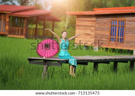 A beautiful young Lao woman wearing a traditional dress sitting on a wooden bridge in a green field happily.