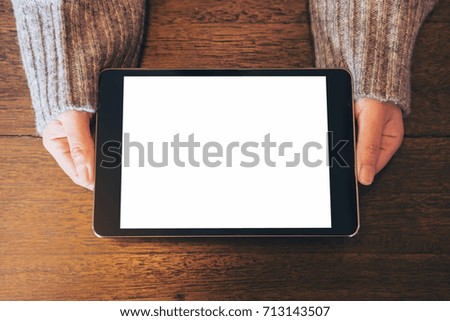 Mockup top view image of woman's hands holding and showing black tablet pc with blank white screen on vintage wooden table 