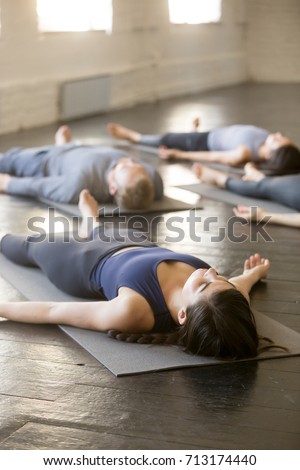 Group of young sporty people practicing yoga lesson with instructor in gym, lying in Dead Body exercise, doing Savasana, Corpse pose, students relaxing after working out in sport club, indoor image  Royalty-Free Stock Photo #713174440