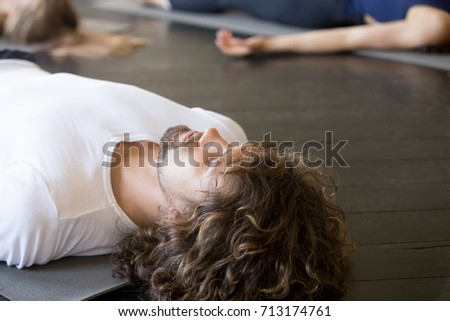 Close up of male head, young handsome man with curly hair, practicing yoga lesson with group in gym, lying in Dead Body exercise, doing Savasana, Corpse pose, relaxing after working out in sport club  Royalty-Free Stock Photo #713174761