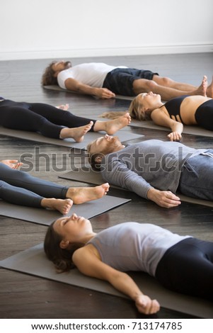 Group of young sporty people practicing yoga lesson with instructor in gym, lying in Dead Body pose, doing Savasana, Corpse exercise, friends relaxing after working out in sport club, indoor image  Royalty-Free Stock Photo #713174785