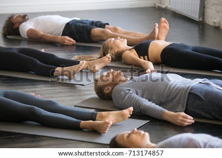 Group of young sporty people practicing yoga lesson with instructor in gym, lying in Dead Body exercise, doing Savasana, Corpse pose, friends relaxing after working out in sport club, indoor image  Royalty-Free Stock Photo #713174857