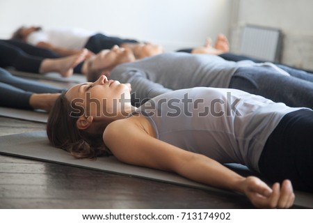 Group of young sporty people practicing yoga lesson with instructor in gym, lying in Dead Body exercise, doing Savasana, Corpse pose, friends relaxing after working out in sport club, studio image 