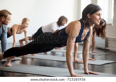 Group of young attractive sporty people practicing yoga lesson with instructor, doing Push ups or press ups exercise, standing in Plank pose, friends working out in club, indoor full length, studio  Royalty-Free Stock Photo #713185936