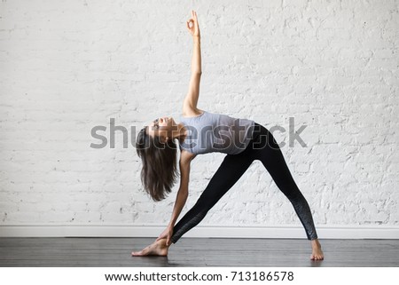 Young woman practicing yoga, standing in Utthita Trikonasana exercise, extended triangle pose, working out, wearing sportswear, gray tank top, black pants, indoor full length, studio background  Royalty-Free Stock Photo #713186578